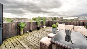 A rooftop patio with wooden decking, potted plants, outdoor furniture, and a view of a cityscape under a cloudy sky.