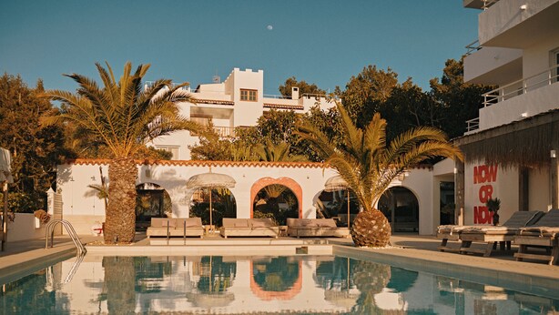 A swimming pool with lounge chairs and palm trees, surrounded by a building with arches and a sign that reads "NOW OR NEVER". - Sunset Oasis Ibiza- Adults Only (Sant Josep de sa Talaia)
