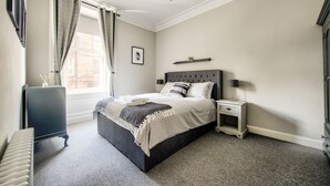 A bedroom with a bed, bedside tables, a radiator, a window with curtains, a chandelier, and a picture on the wall. - Stylish Modern Apartment Parking (Edinburgh)