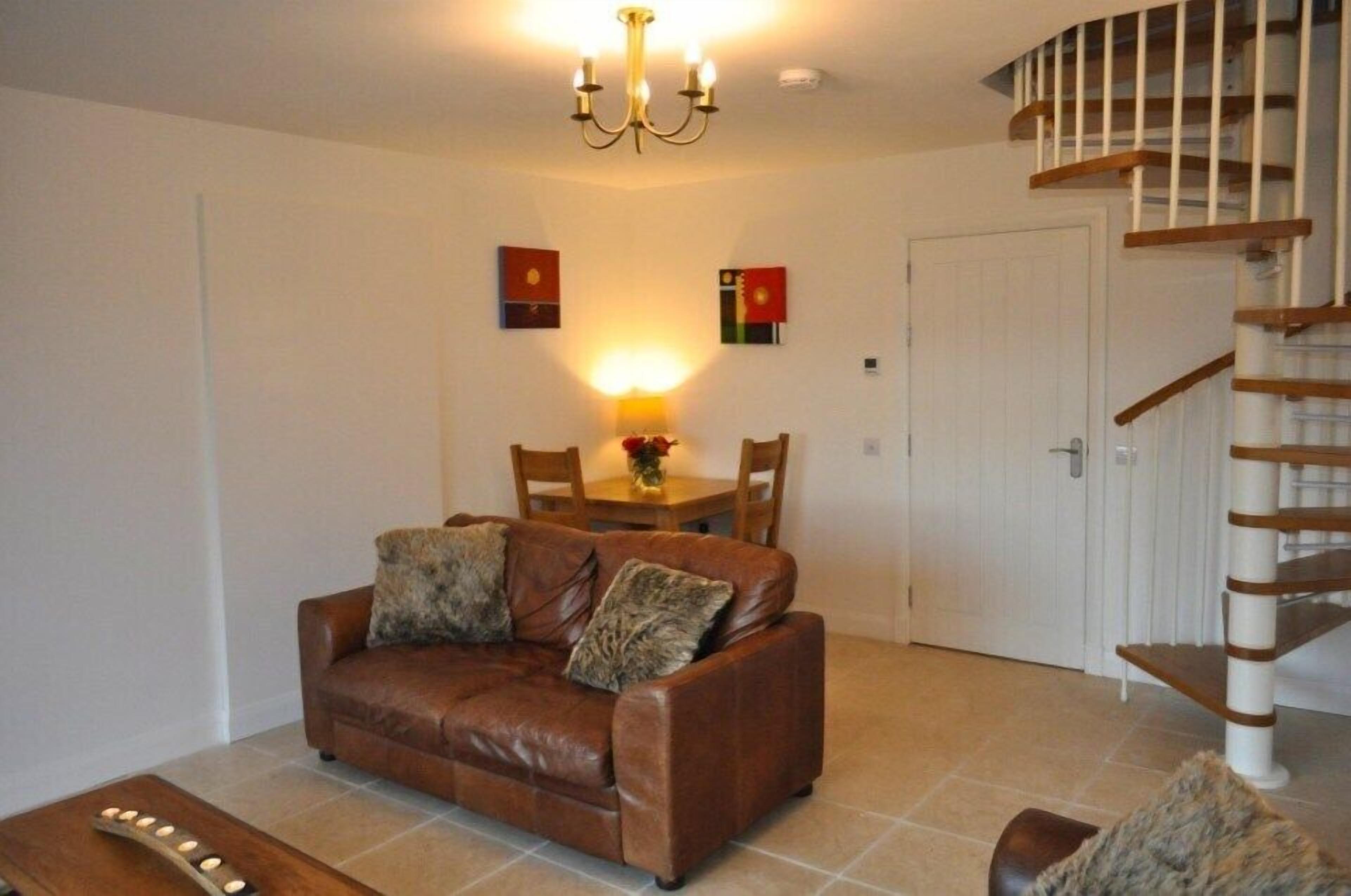 A living room with a brown leather sofa, a wooden coffee table, a chandelier, a dining table with chairs, and a staircase.