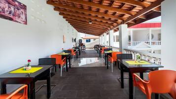 Un espace extérieur abrité, aménagé pour les repas, avec des tables et des chaises orange, un plafond en bois et une vue sur un bâtiment en arrière-plan.