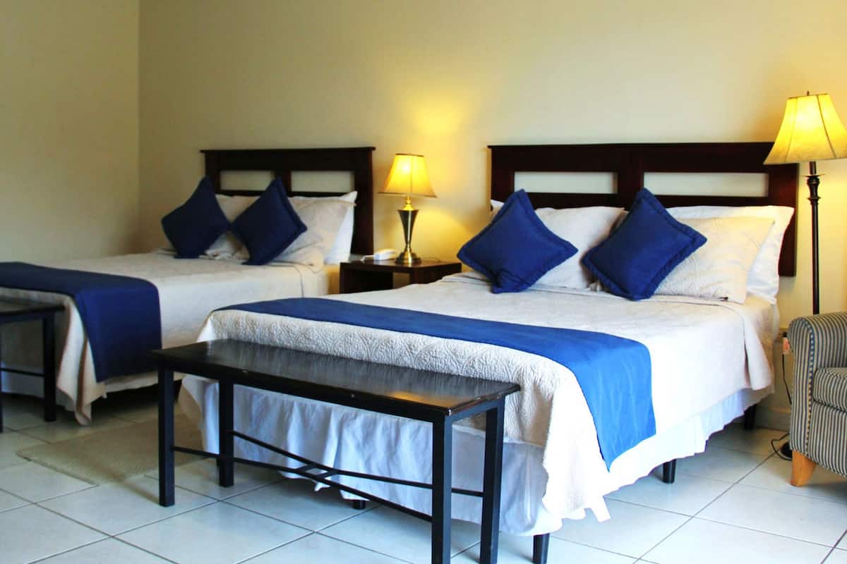 A hotel room with two beds, each with blue and white bedding, a small table, and a lamp on a nightstand.