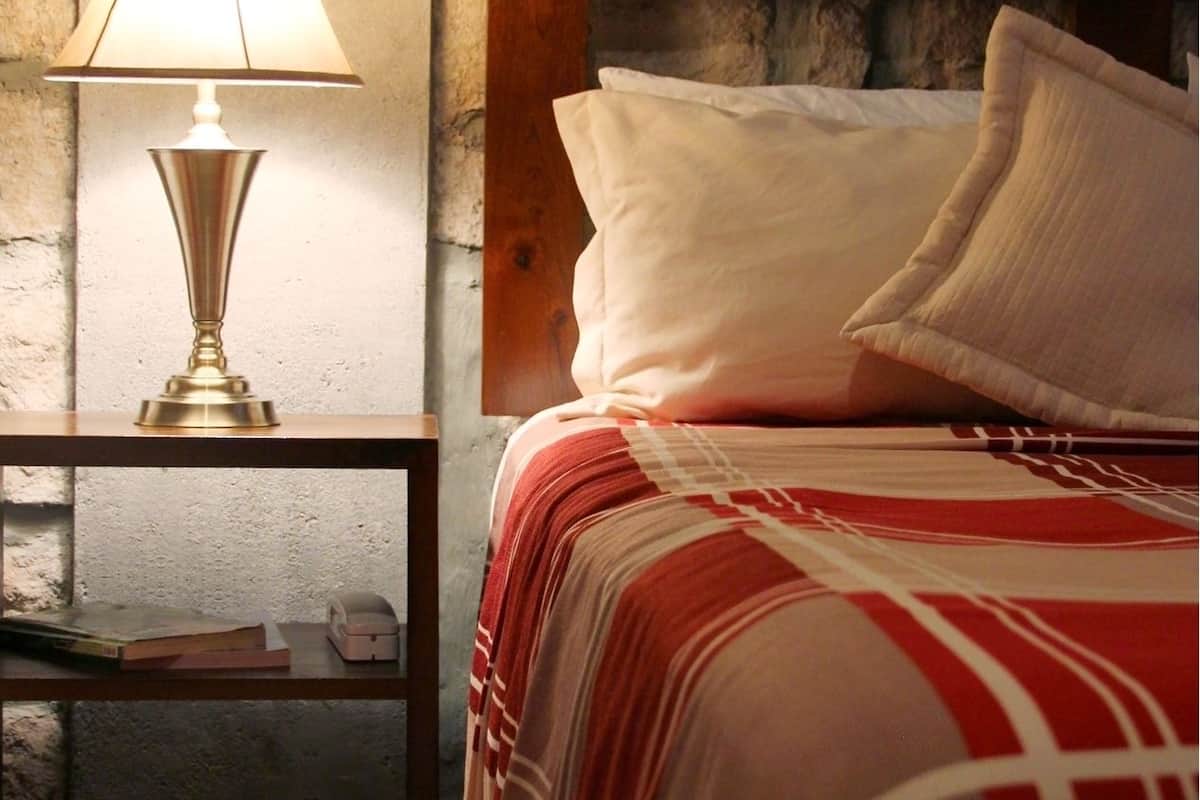 A bedroom with a stone wall, a wooden headboard, a bedside table with a lamp, and a bed with a red and white striped bedspread.