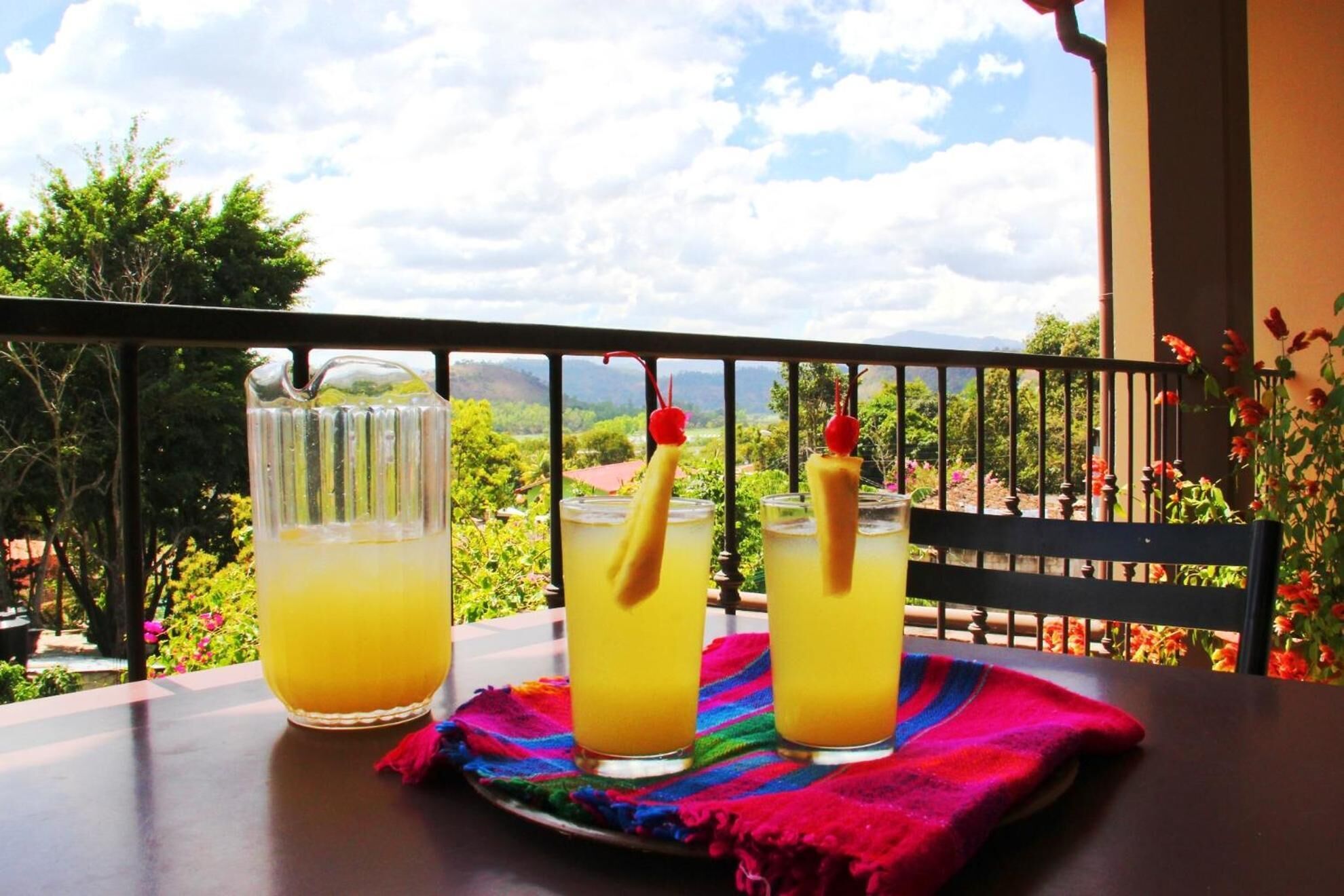 Three glasses of orange juice on a table with a colorful cloth, set against a balcony railing overlooking a lush garden and distant mountains.