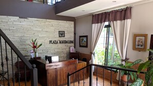 Un mostrador de recepción con un fondo de pared de piedra, una escalera y plantas en macetas.