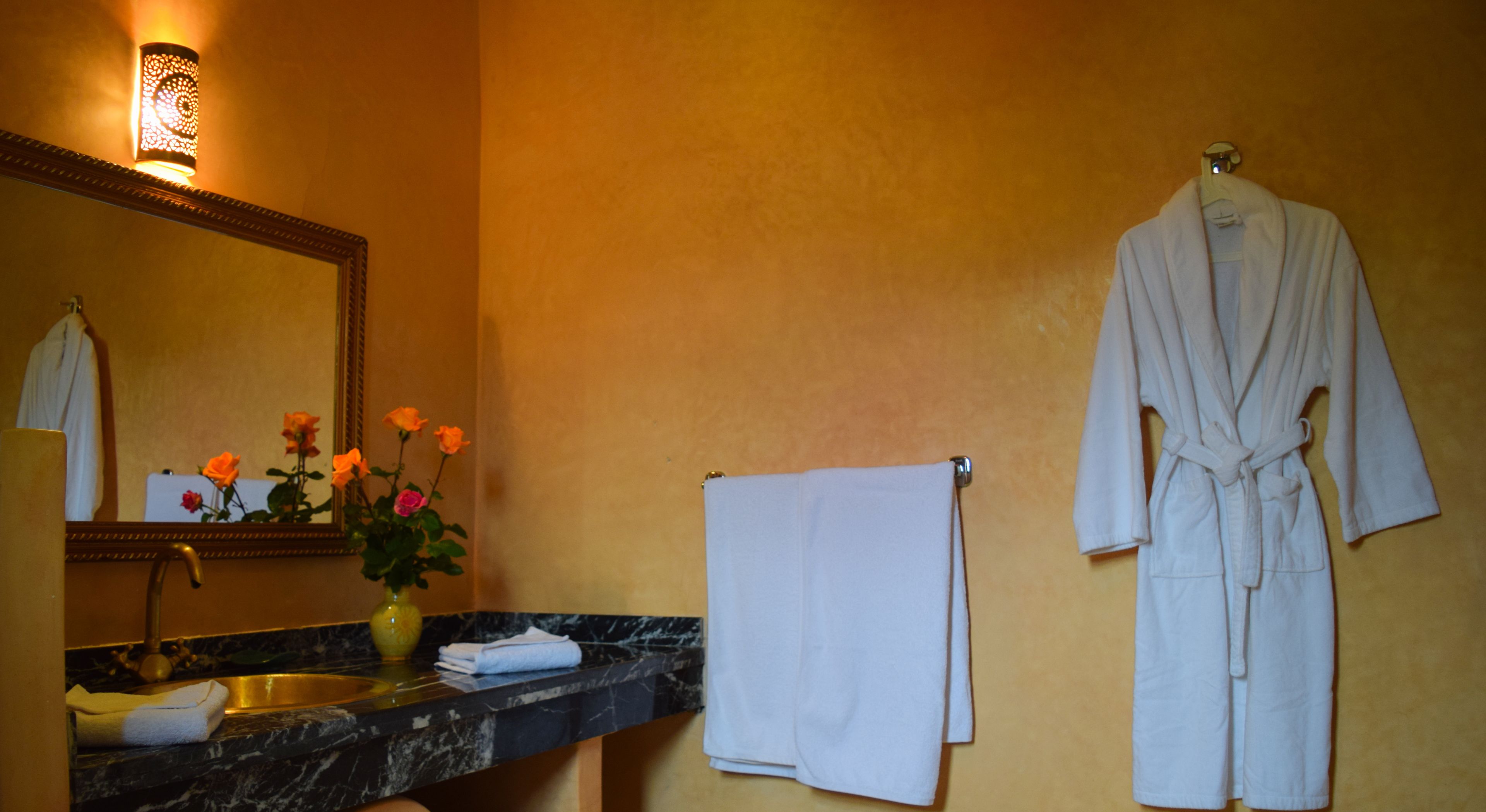 A bathroom with a marble sink, a mirror, a vase with flowers, a white robe, and a towel rack.