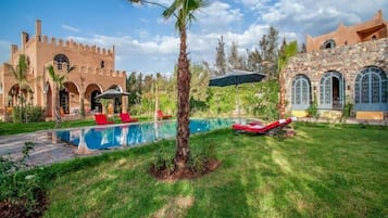 Un área de piscina con una palmera, sillas de descanso y un edificio de piedra con arcos.