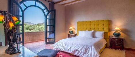 A bedroom with a yellow headboard, two bedside lamps, a red patterned rug, and a view of the outdoors through a large arched window.