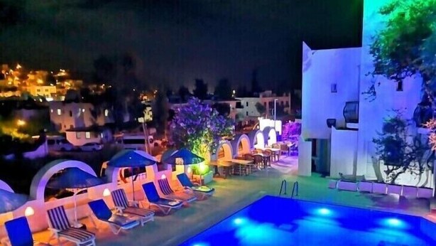 Un espace piscine doté de chaises longues et de parasols, entouré de bâtiments illuminés.