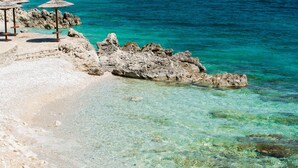 Una costa rocosa con agua turquesa cristalina, un sendero de piedra y algunas sombrillas.