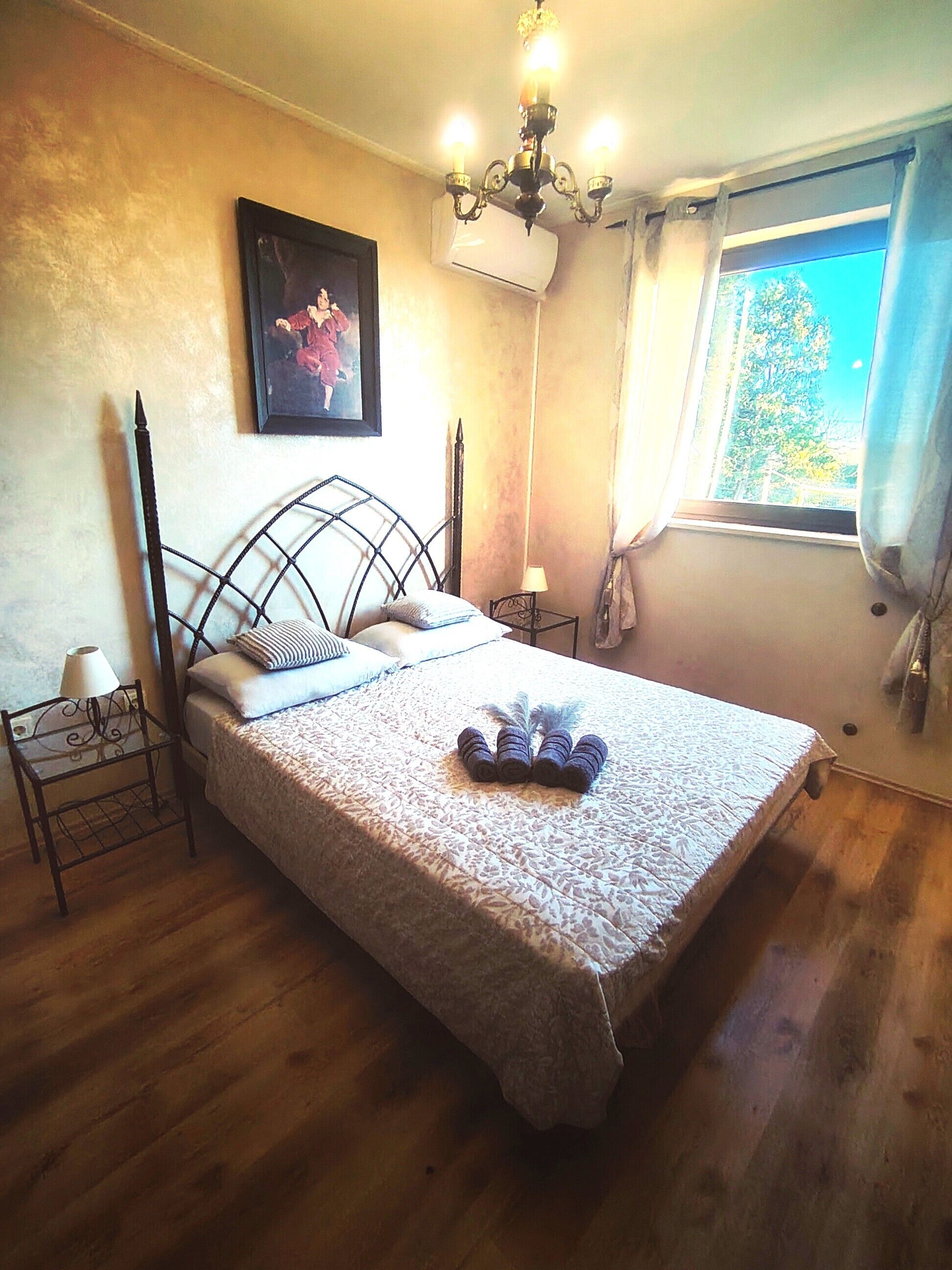 A bedroom with a bed, a nightstand, a lamp, a painting, an air conditioning unit, and a window with curtains.