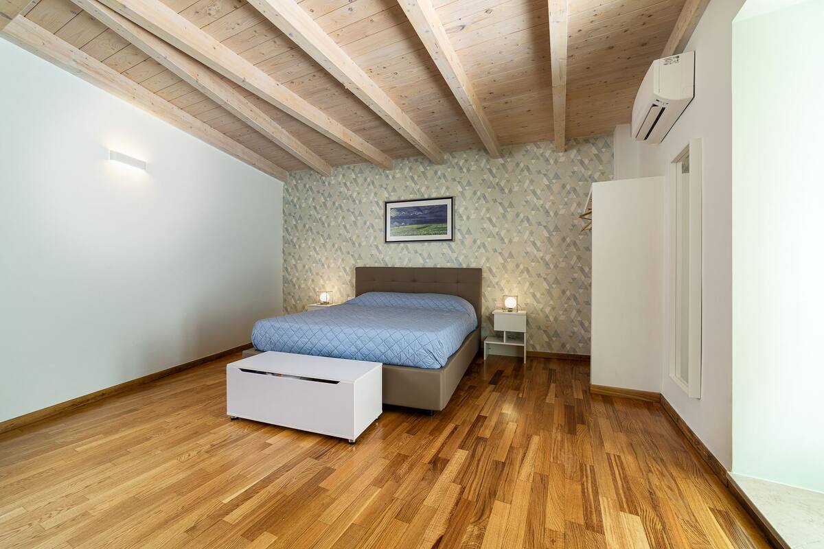Une chambre avec un plafond en bois, un lit recouvert d’une couverture bleue, deux tables de chevet avec des lampes et une petite armoire blanche.