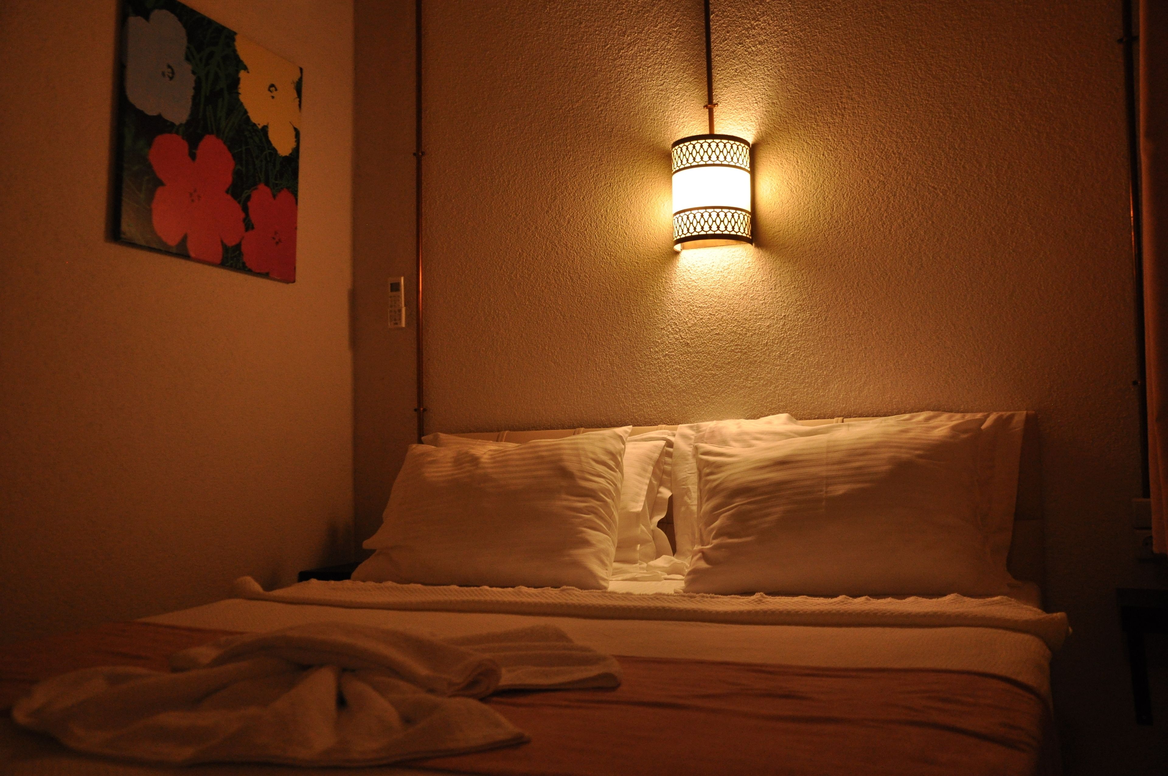 A dimly lit hotel room with a bed, pillows, and a wall-mounted light fixture.