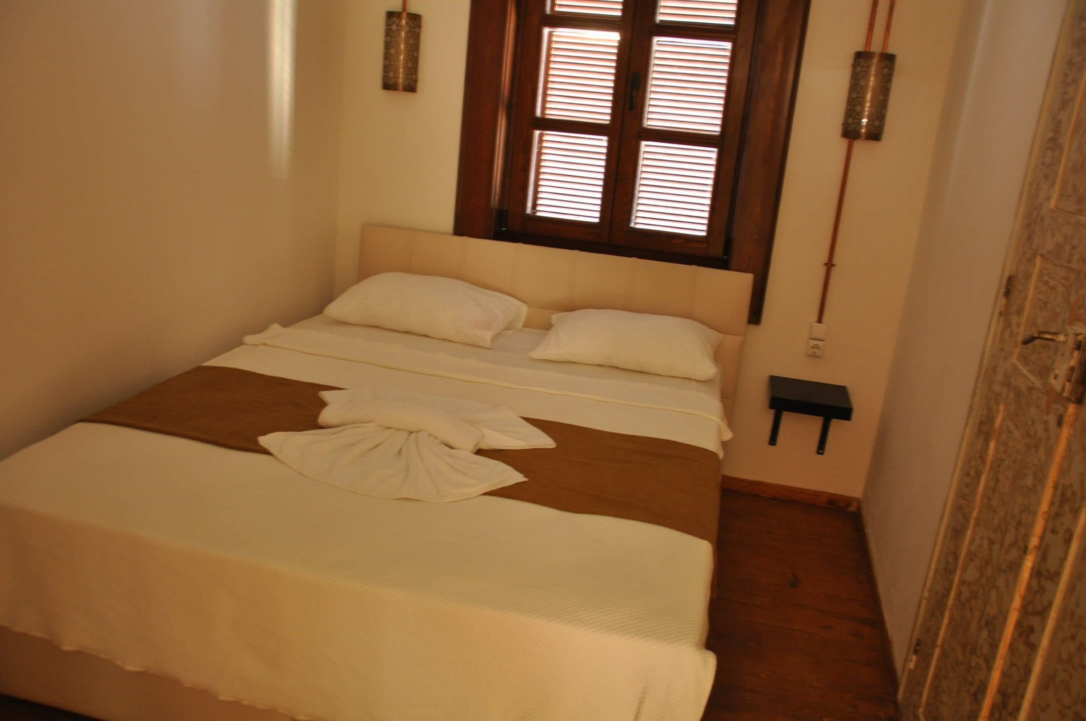 Una cama bien hecha con una manta marrón y almohadas blancas, una mesita de noche y una ventana con contraventanas de madera.