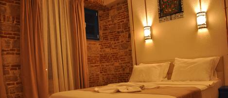 A bed with white linens and a brown blanket, a wooden floor, a brick wall, and a window with blue shutters.