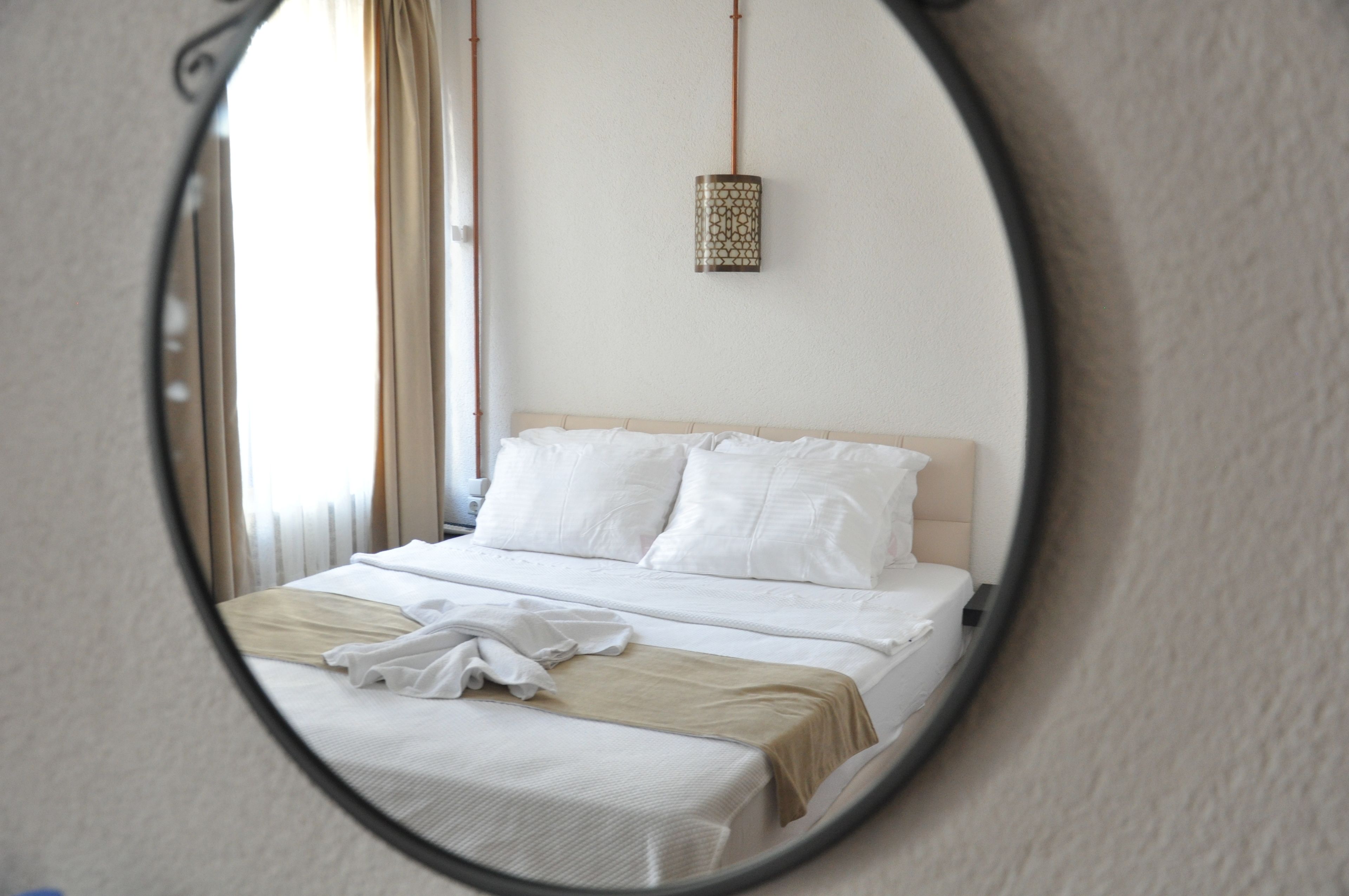 Un lit bien fait, avec du linge de lit blanc et un plaid beige, un luminaire suspendu et une fenêtre avec des rideaux.