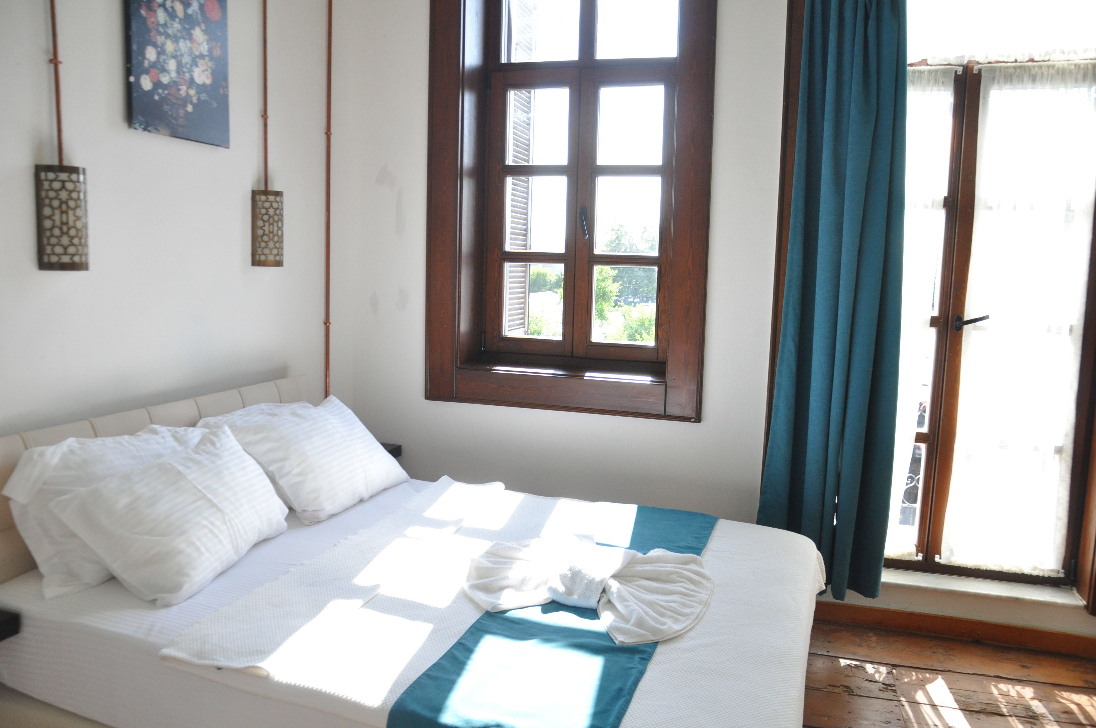 Una cama con ropa de cama blanca y un detalle en azul, una ventana con vistas a la vegetación y dos lámparas fijadas a la pared.