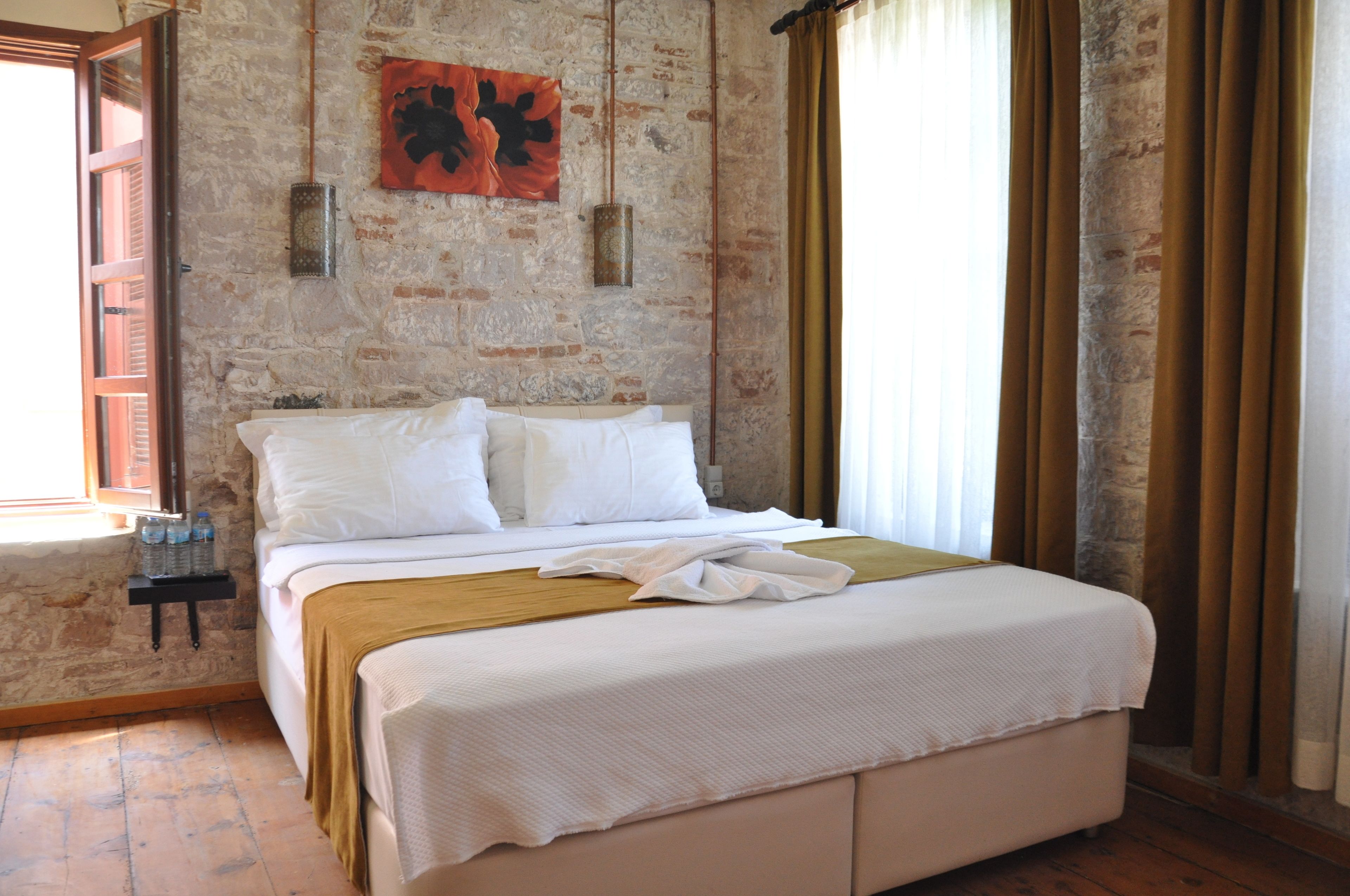 Un dormitorio con pared de piedra, una cama con ropa de cama blanca, una mesita de noche pequeña con botellas y una ventana con cortinas.