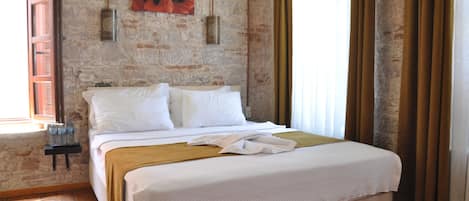 Un dormitorio con pared de piedra, una cama con ropa de cama blanca, una mesita de noche pequeña con botellas y una ventana con cortinas.