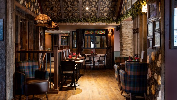 A cozy, rustic interior with wooden floors, plaid-patterned chairs, and a dining area with a stone wall and hanging lights. - The Ambleside Inn - The Inn Collection Group (Ambleside)
