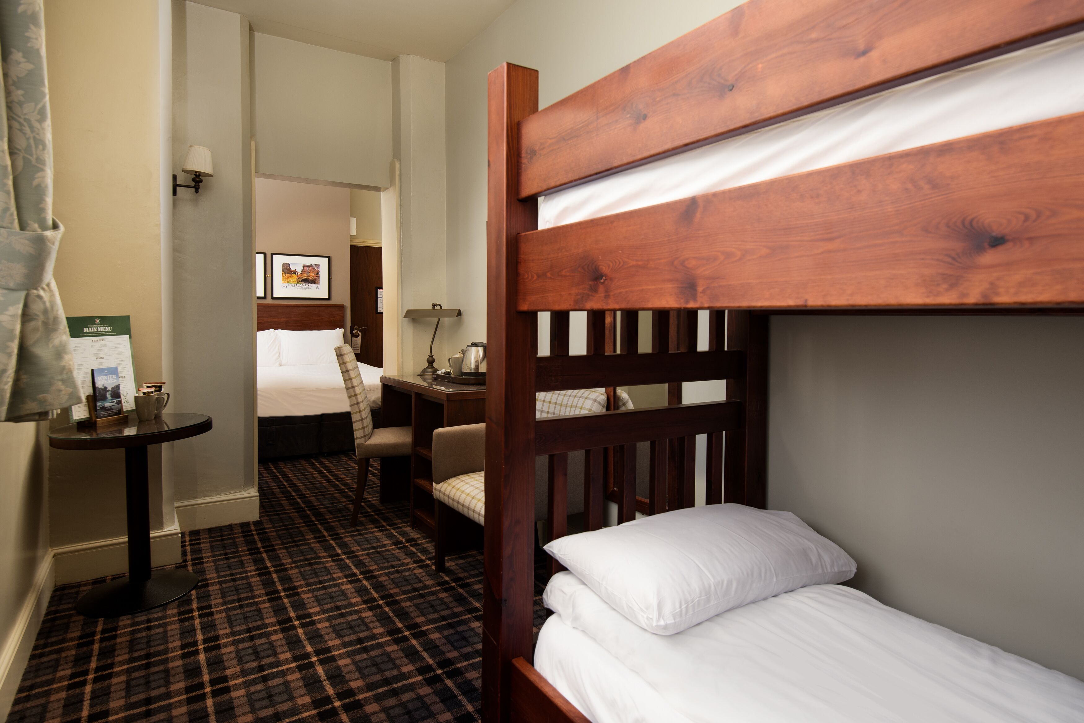 A hotel room with a bunk bed, a desk with a chair, a small table with a lamp, and a check-in counter.