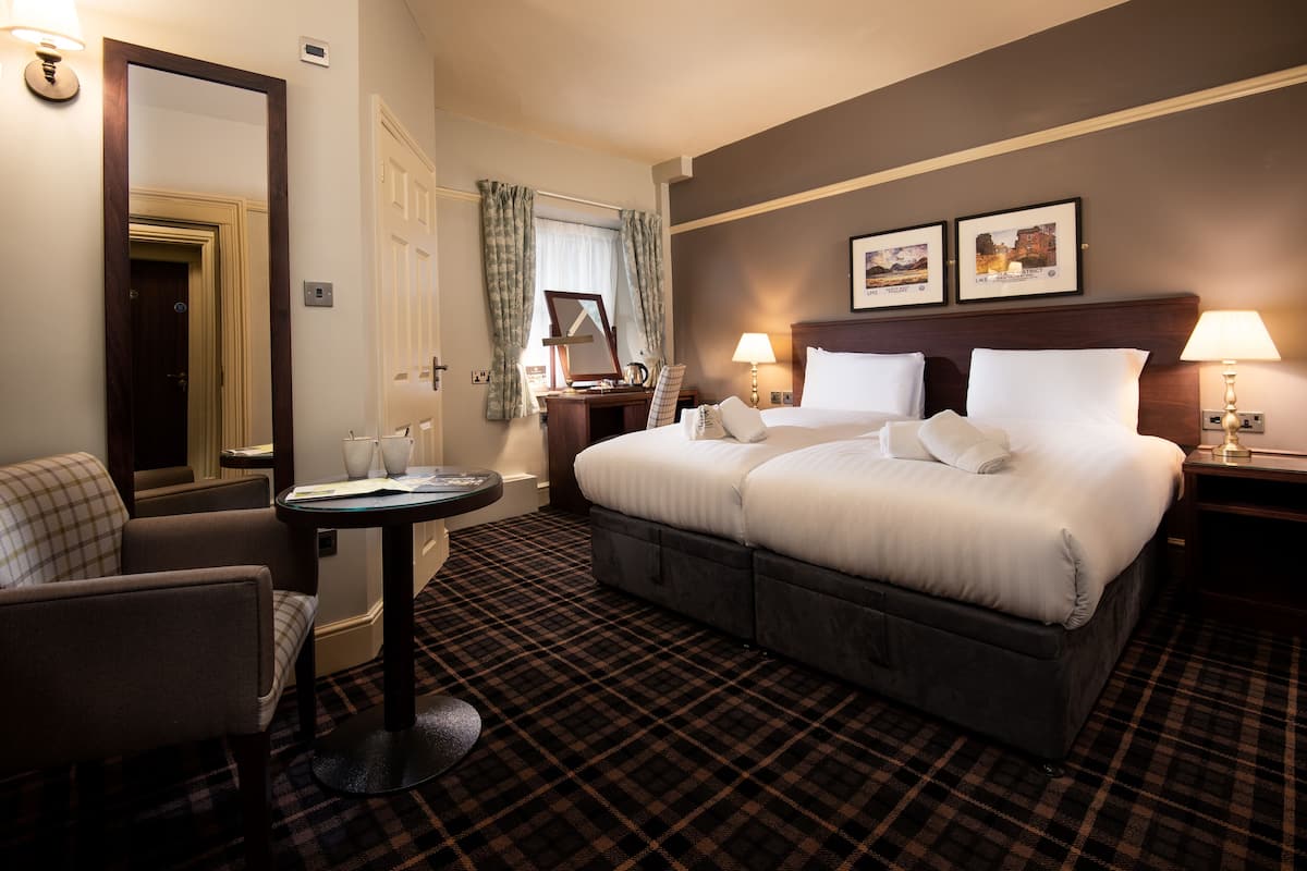 A hotel room with a bed, a chair, a table, a mirror, and a wall with two framed pictures.