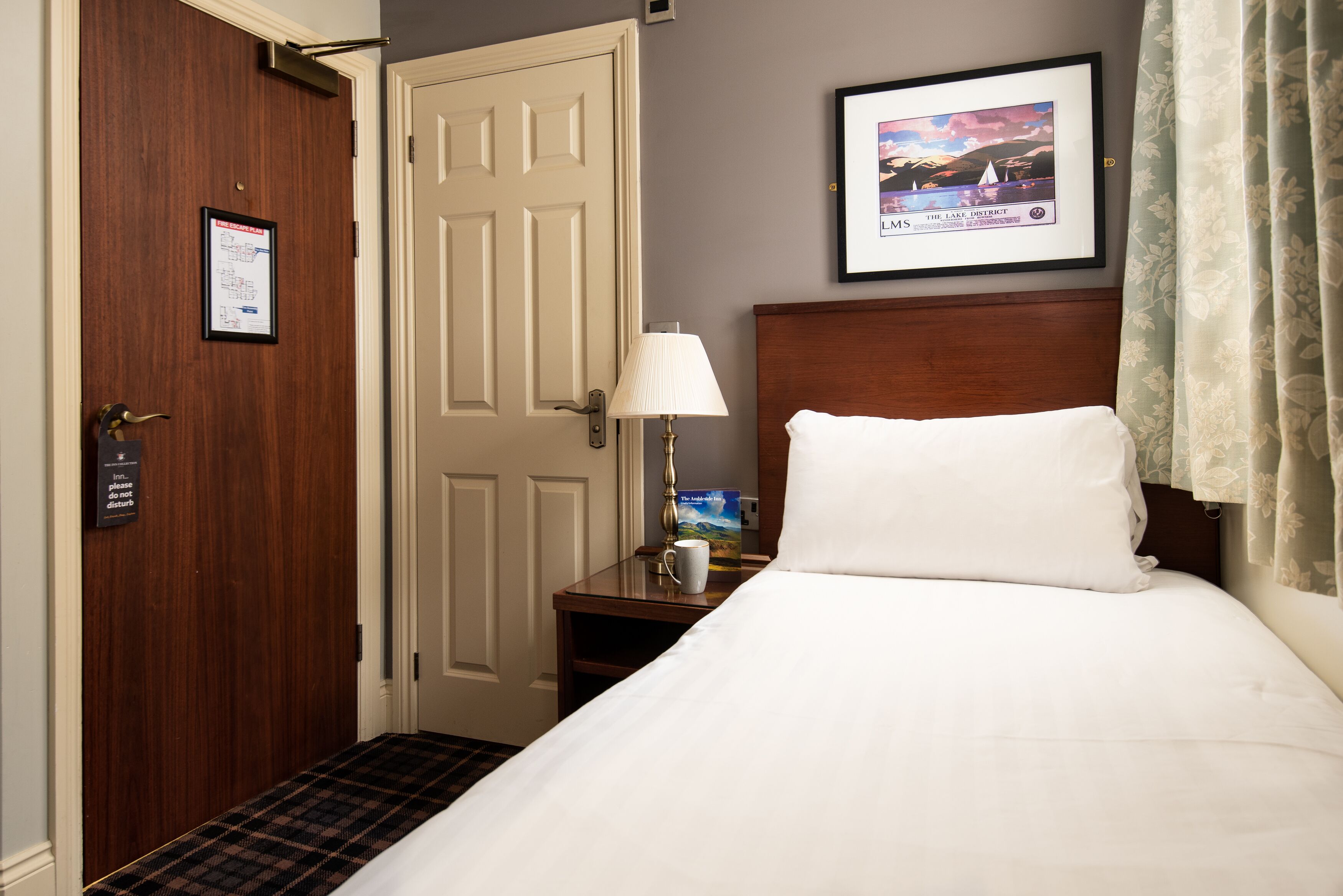 Een hotelkamer met een bed, een nachtkastje met lamp, een lijst met afbeelding en een deur met een bordje.