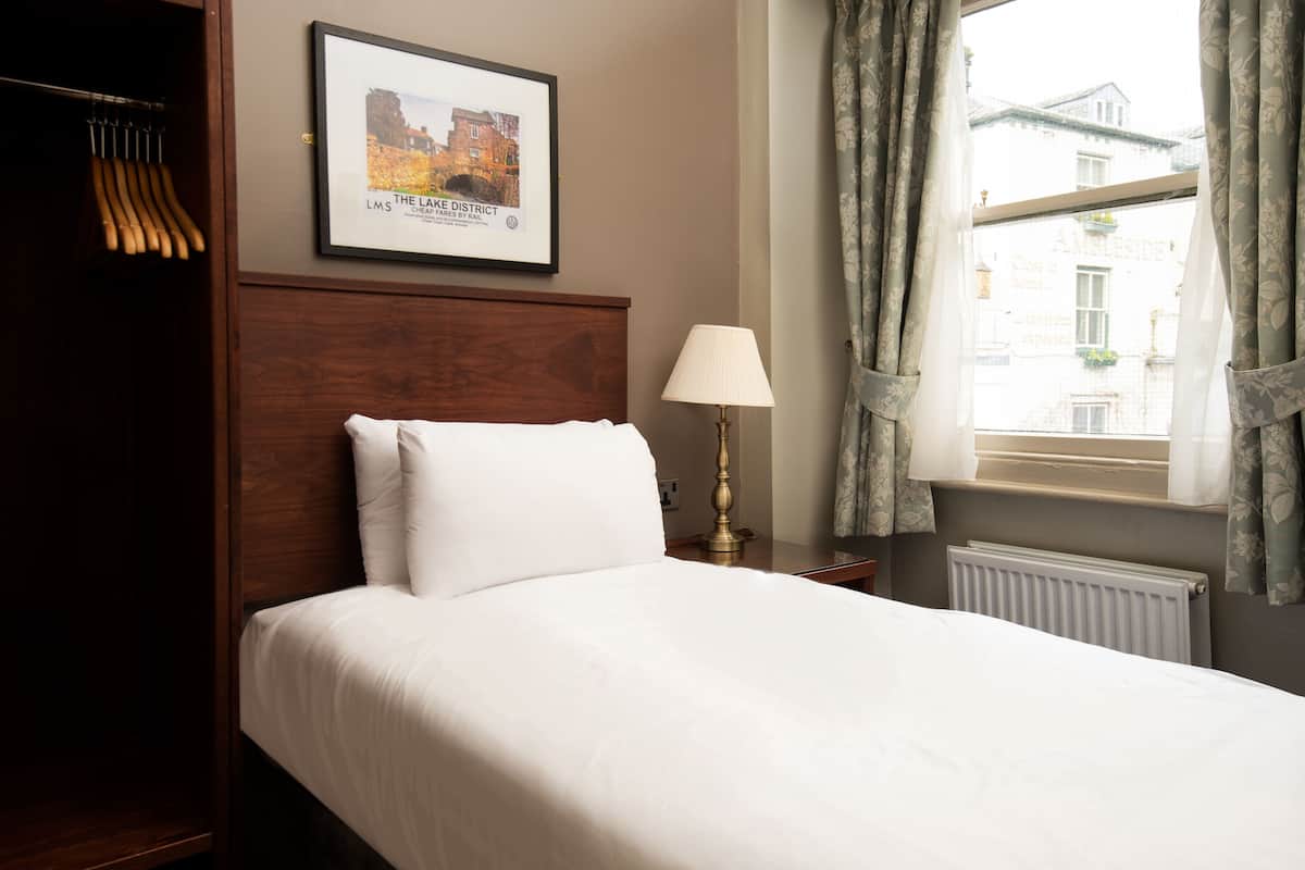 A neatly made bed with white linens, a framed picture of a building on the wall, a lamp on a bedside table, and a window with curtains.