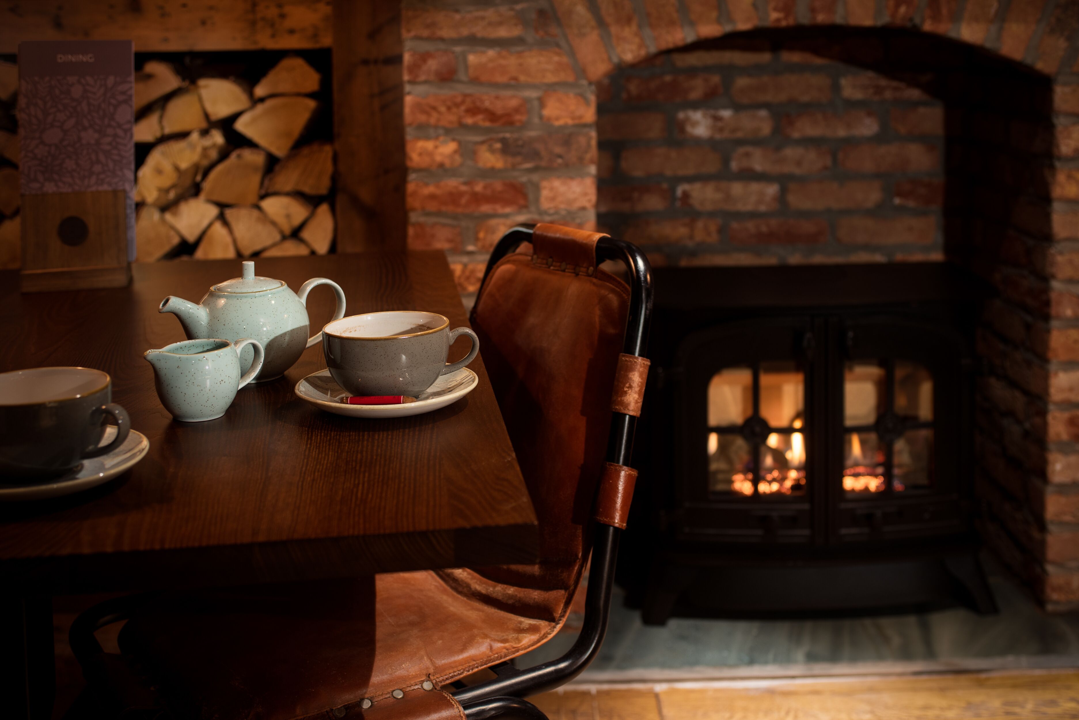 A wooden dining table with a teapot, cups, and saucers. A leather chair is positioned in front of the table. A brick fireplace with a fire is visible in the background.