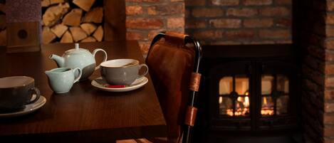A wooden dining table with a teapot, cups, and saucers. A leather chair is positioned in front of the table. A brick fireplace with a fire is visible in the background.