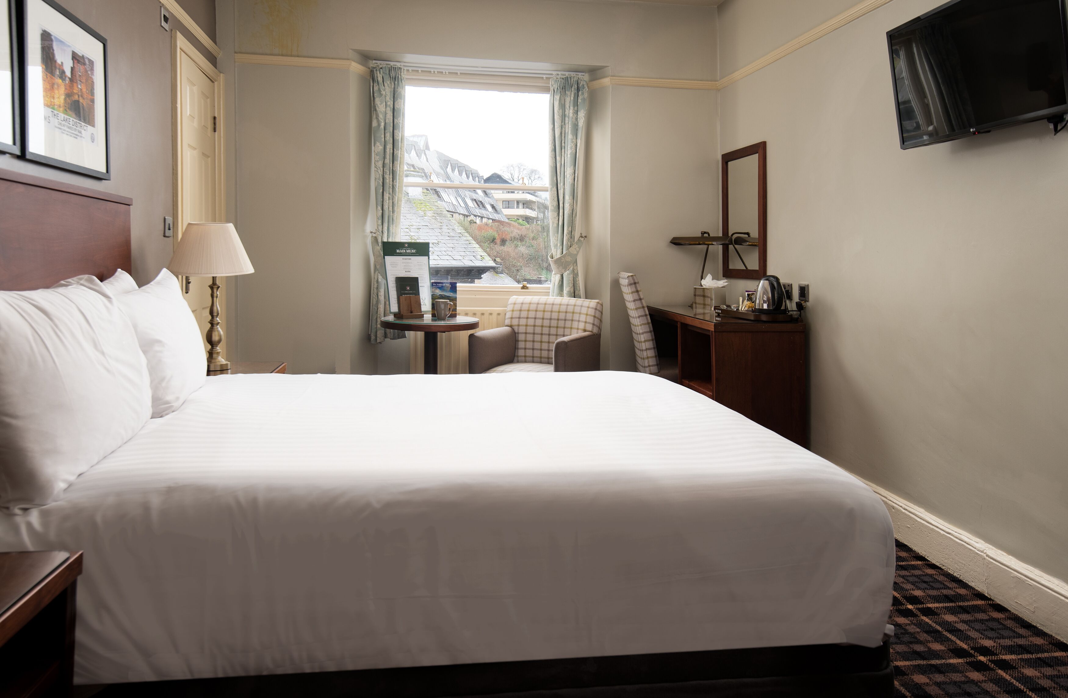 A hotel room with a large bed, a desk with a chair, a small table with a lamp, a television, and a window with a view of buildings.