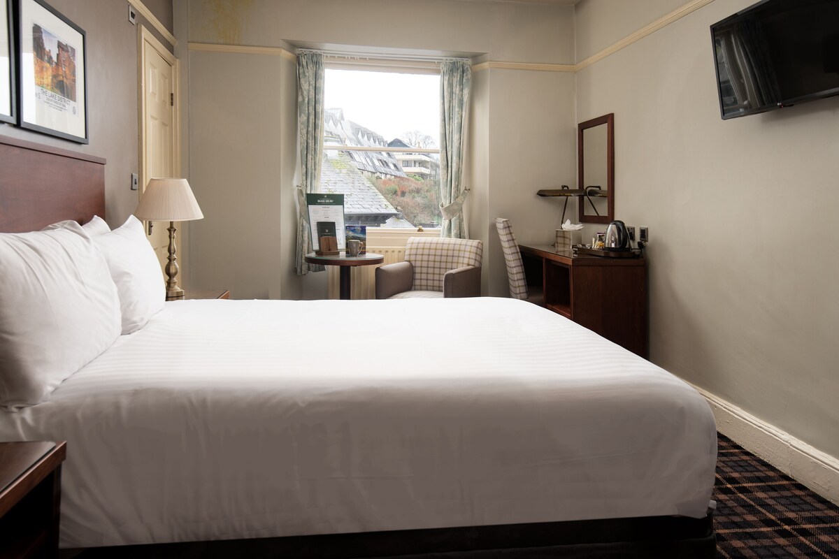A hotel room with a large bed, a desk with a chair, a small table with a lamp, a television, and a window with a view of buildings.