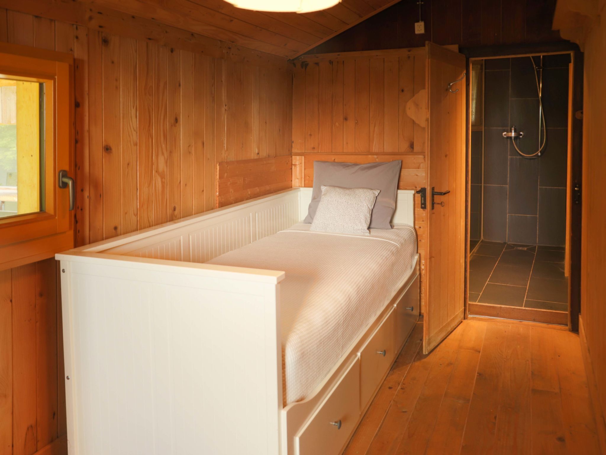 A wooden cabin room with a bed, a small window, and a shower area.