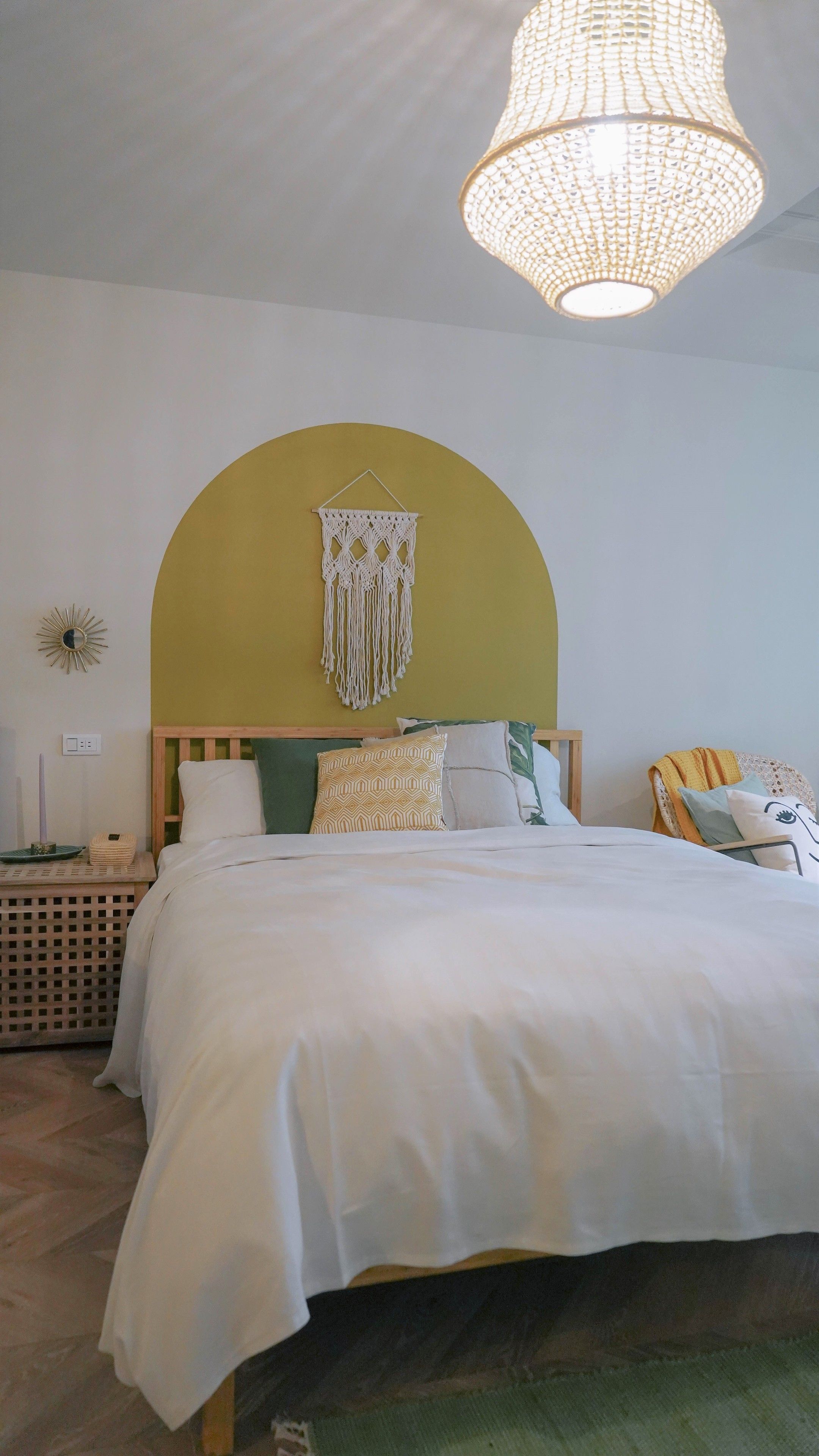 A bedroom with a bed, a hanging macrame wall decoration, a wooden headboard, and a hanging light fixture.