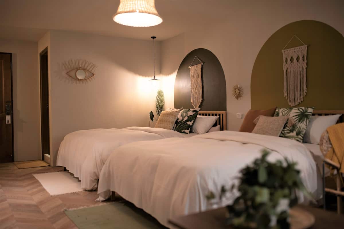 Un dormitorio con dos camas, una lámpara colgante, un tapiz de macramé y un espejo decorativo.