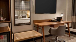 Une chambre d’hôtel avec un bureau en bois, une chaise, un lit avec une tête de lit, un grand miroir et un téléviseur à écran plat.
