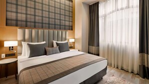 Une chambre d’hôtel avec un grand lit, deux tables de chevet avec des lampes, une tête de lit à motifs géométriques et des rideaux légers à la fenêtre.