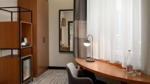 Une chambre d’hôtel avec un bureau en bois, une chaise, un miroir, une lampe, un mini-bar et une grande fenêtre avec des rideaux légers.