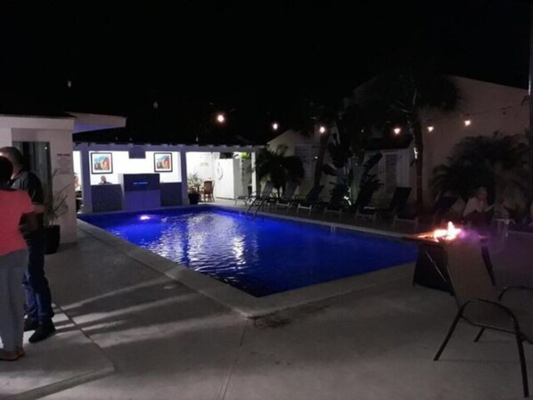 Un espace piscine nocturne avec une piscine illuminée de lumière bleue, des chaises longues et un petit bâtiment où sont exposées des œuvres d’art.