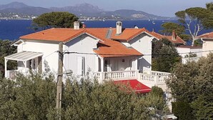 A white building with a red roof, surrounded by greenery and overlooking a body of water with boats. - Small 30m2 house for 2 people on private property with swimming pool (Fréjus)