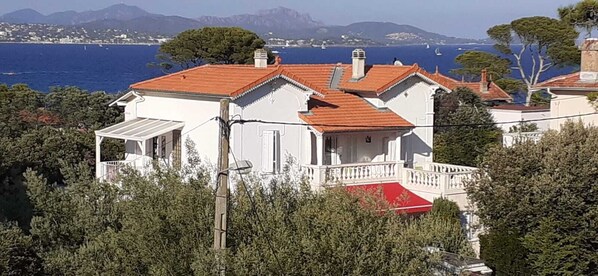 A white building with a red roof, surrounded by greenery and overlooking a body of water with boats. - Small 30m2 house for 2 people on private property with swimming pool (Fréjus)