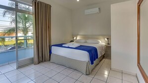 A hotel room with a bed, a ceiling fan, a window with curtains, and a view of palm trees. - Marazul Apart Hotel (Florianópolis)