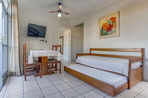 A room with a wooden bed, a table with a tablecloth, a chair, a ceiling fan, a TV mounted on the wall, and a painting of flowers.