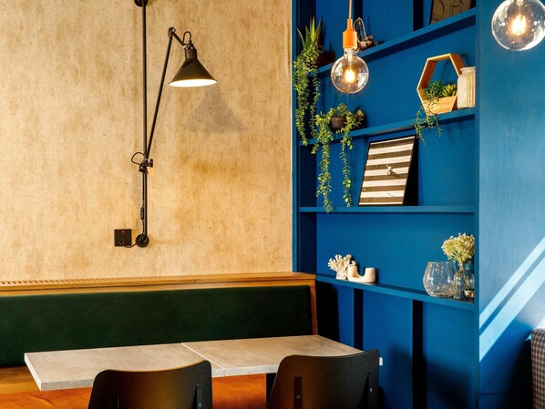 A restaurant interior with a long table, black chairs, a blue bookshelf with plants, and a hanging light fixture. - ibis Wakefield East-Castleford (Castleford)