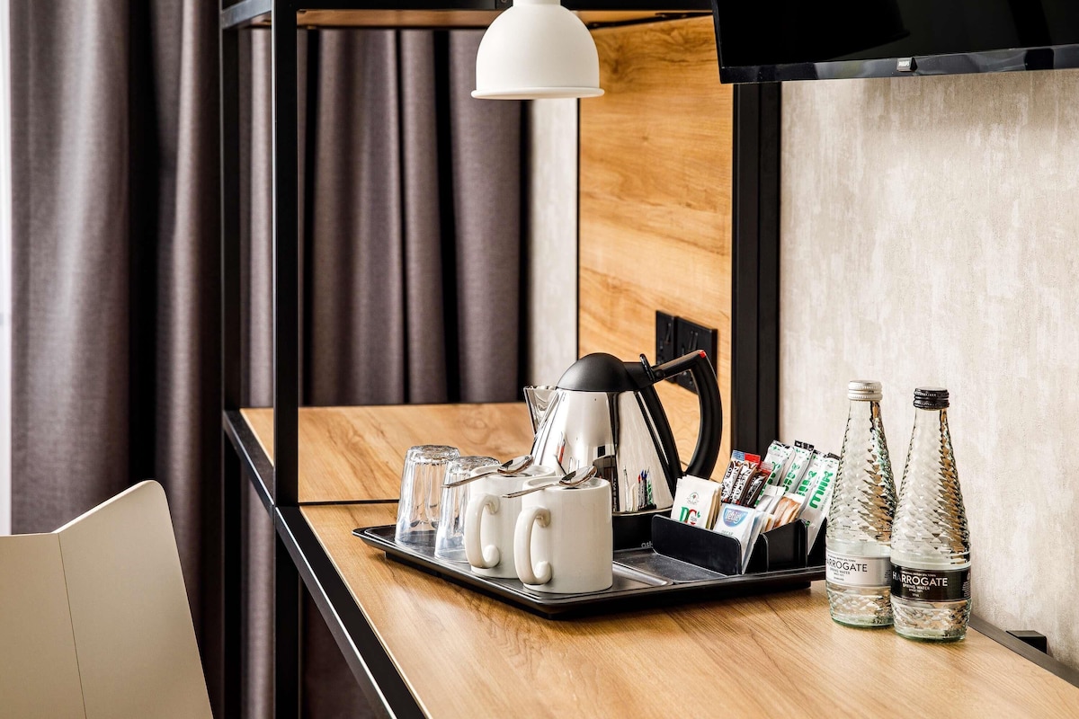 A hotel room with a wooden desk, a kettle, glasses, a tray, and two Harrogate water bottles.