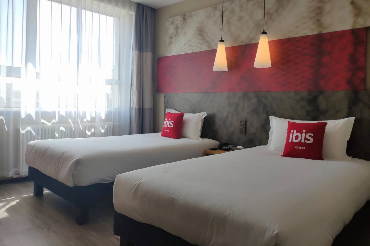 Een hotelkamer met twee bedden, een groot raam met dunne gordijnen en een wand met een marmerlook.