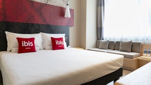 A hotel room with a large bed, two red pillows with "ibis Hotels" text, a bedside table with a lamp, and a window with curtains.