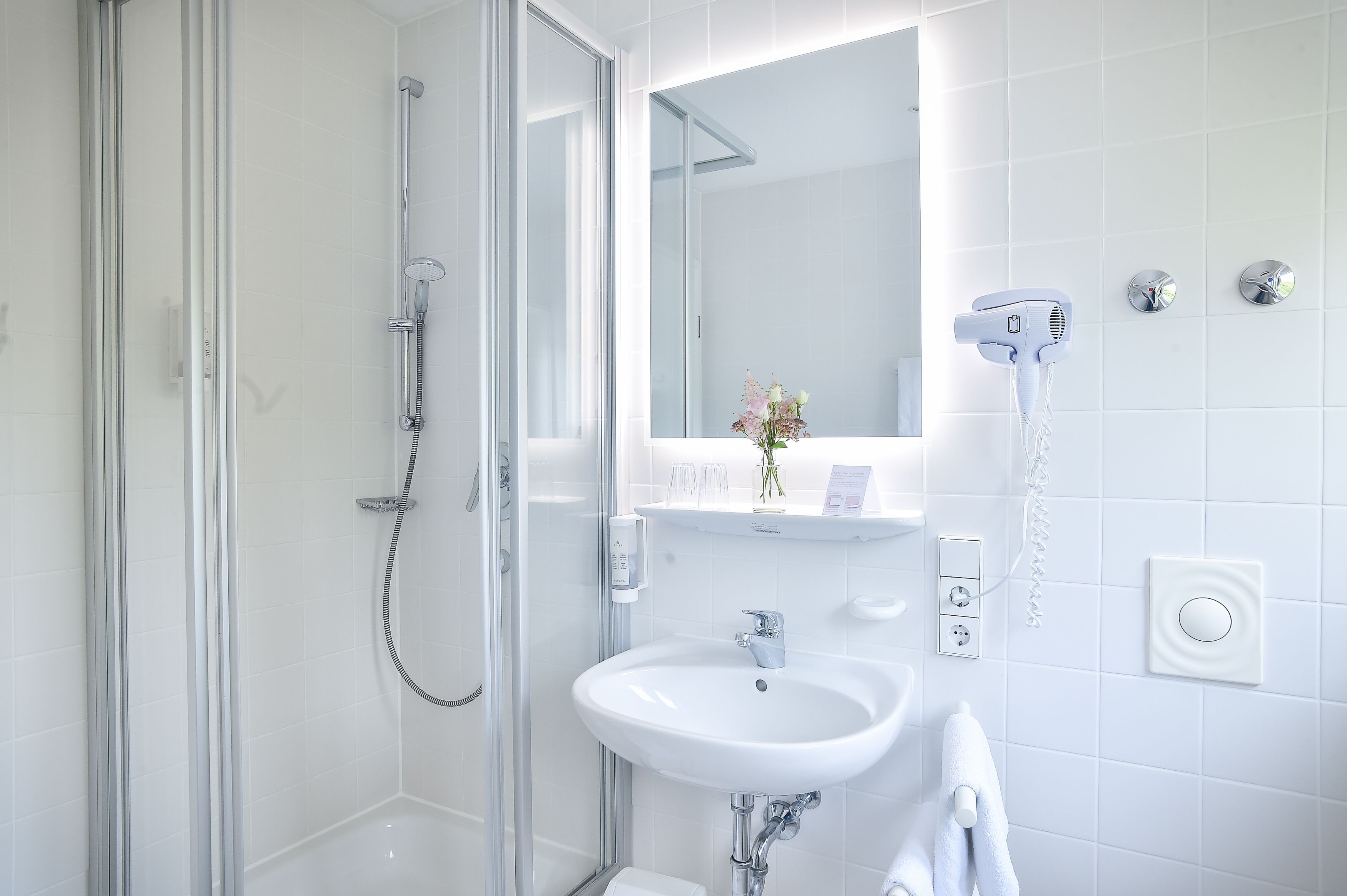 Een moderne badkamer met een douchecabine van glas, een witte wastafel en een haardroger bevestigd aan de muur.