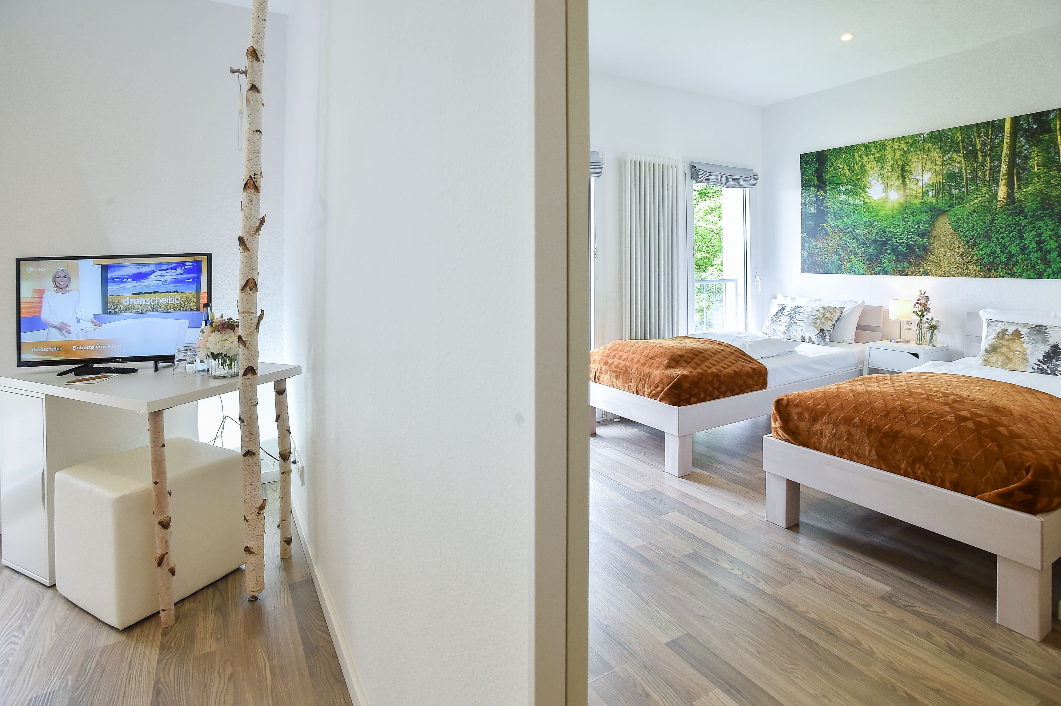 A modern hotel room with a large bed, a smaller bed, a desk with a computer, and a large forest-themed wall mural.