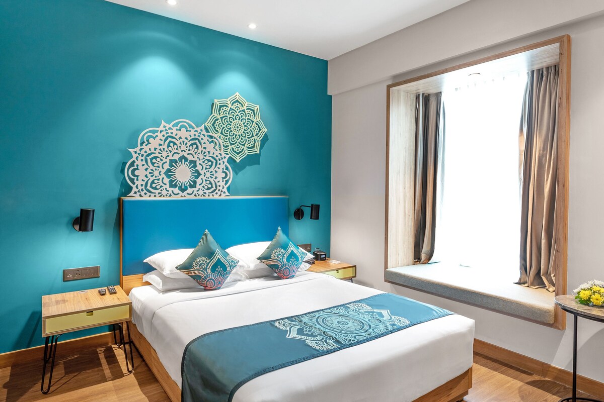 A modern bedroom with a teal accent wall, a large bed with patterned pillows, a bedside table, and a window with curtains.